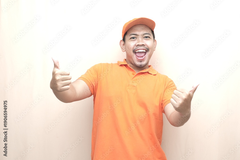 Naklejka premium Southeast Asian man in orange uniform and cap showing two thumbs up on beige background, expressing approval, success, and service satisfaction.