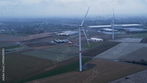 Aerial view of wind turbines standing tall in a landscape of fields and industrial warehouses, with a muted color palette, Tournai, Wallonia, Belgium.