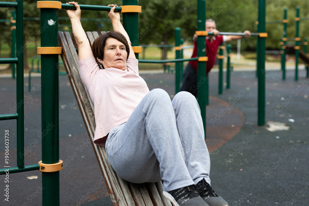 Fototapeta premium Mature woman goes in for sports in city park on sports ground - doing exercises to pull his legs to chest