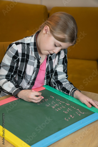 Young schoolgirl learning and completing math homework, writing arithmetic examples on a green framed chalkboard with pink chalk, focusing on her studies at home. Vertical photo