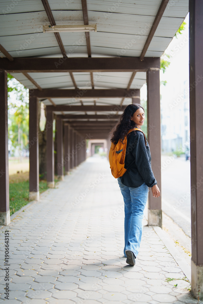 Fototapeta premium Young woman with backpack walking along a covered outdoor pathway, pausing and looking back, conveying concepts of journey, discovery, and urban exploration