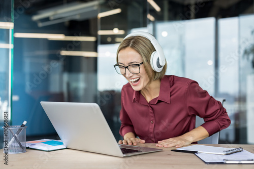 Young businesswoman wearing headphones and eyeglasses laughing sincerely while looking at her laptop screen, enjoying positive news or entertaining content during work in a modern office