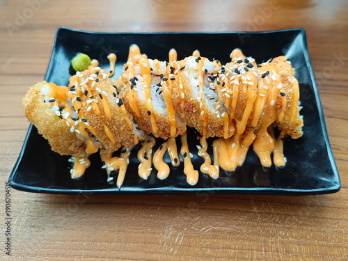 Crispy fried sushi roll served on a black plate, topped with spicy mayonnaise sauce and sesame seeds on a wooden table.