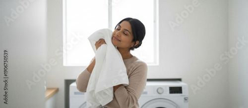 Indian woman smelling fresh laundry and holding white towel in laundry room with washing machine