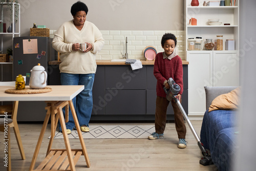 Tableau sur toile Black woman standing in kitchen holding mug watching Black boy vacuuming floor w