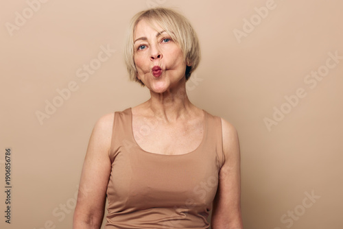 Schilderij op canvas Mature woman with short blonde hair wearing beige tank top makes playful face with puckered lips on neutral background