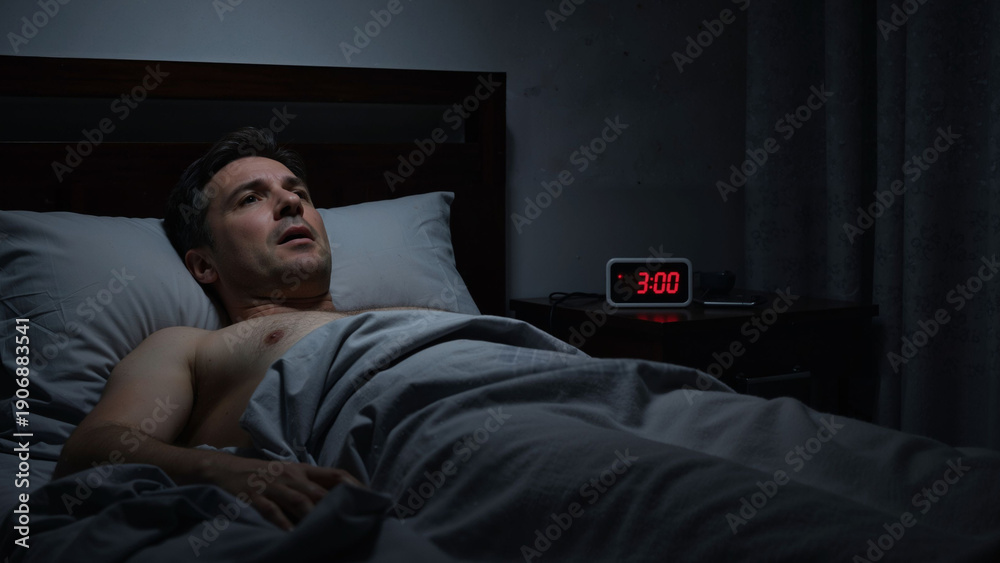 Obraz premium Caucasian man experiencing insomnia late at night, unable to sleep with an alarm clock showing 3:00 am next to his bed. Sleep disorder concept.