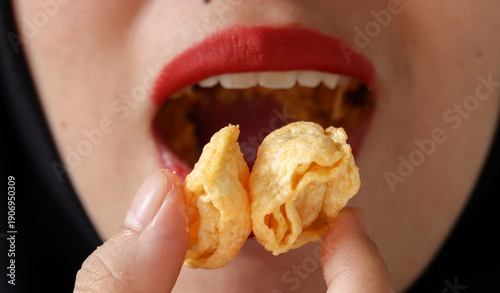 Fotografi Close up of an Asian Muslim woman wearing hijab and red lipstick eating a crunchy corn snack, focusing on texture, snack craving, bold makeup, and modern lifestyle concept