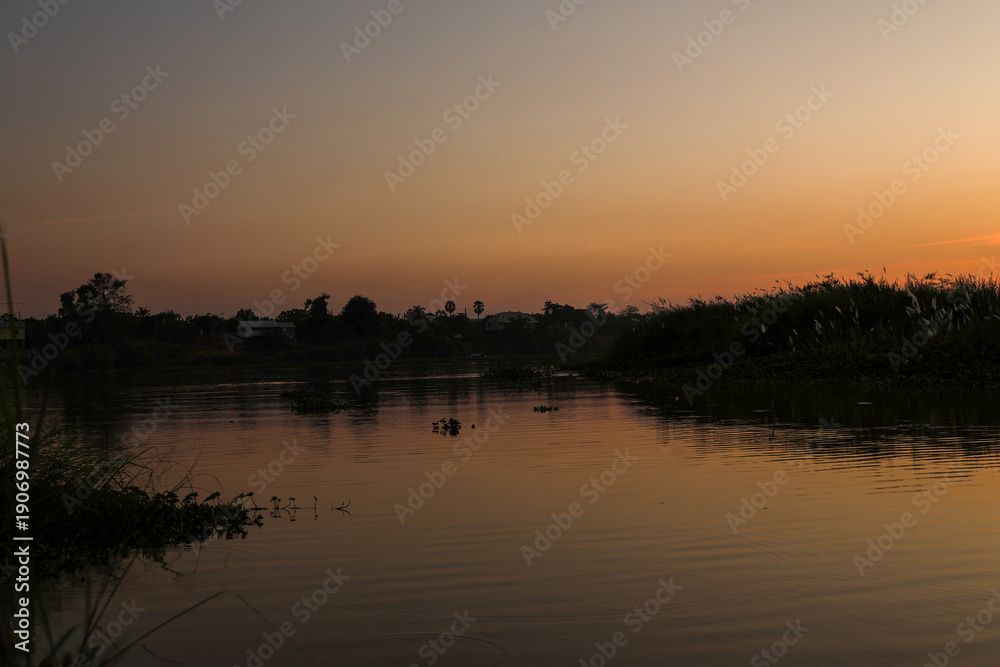 Fototapeta premium Quiet water surface reflecting dramatic golden hour sunset light over dark tropical forest and rural homes, creating a peaceful, tranquil atmosphere.