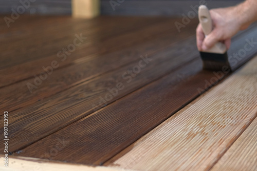 Brown oil for protection of wood by staining. Painting a larch for protecting and pests. Woodworker applying finish to terrace deck board. Repairman varnishing with brush wooden plank floor surface.
