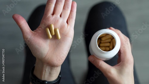 Top View of Woman Pouring Supplement Capsules Into Palm. A woman pours yellow medicine capsules from a white plastic bottle into her hand. Healthy habits, pharmaceutical treatment, and vitamin intake