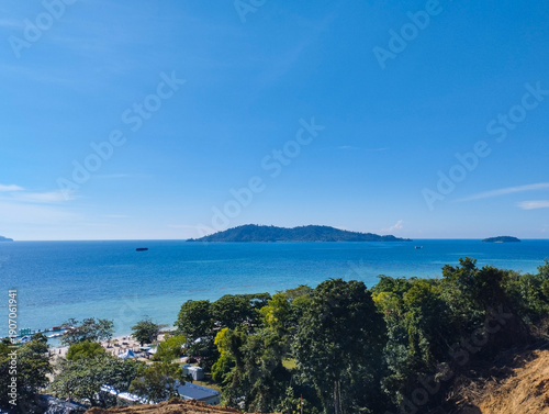 A high-angle view of a lush coastline overlooking a calm turquoise sea and a distant mountain island under a clear blue sky.