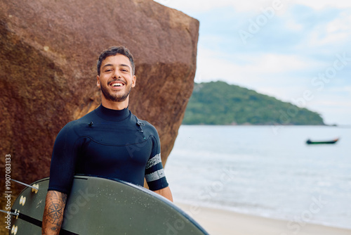 Happy young man wearing a wetsuit and holding a surfboard, standing on a sandy beach next to a large rock