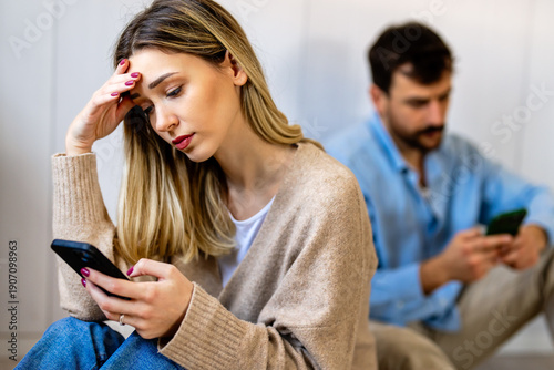 Fotografija Young couple sitting back to back, both focused on their smartphones, appearing