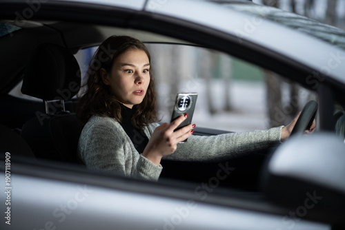Fotografie young girl looks at her smartphone with a strong expression of surprise while driving, distracted by a shocking message, notification, or GPS update, creating a dangerous situation