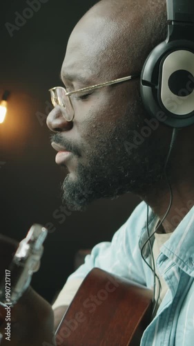 Vertical shot of African American male musician wearing headphones performing guitar melody and vocalizing into microphone while producing song in home studio