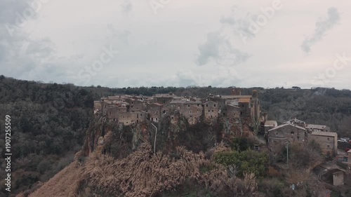 Aerial view of Calcata Vecchia, an ancient medieval village perched on a rocky cliff in Lazio, during a gloomy day. Historic stone houses surrounded by forest and dramatic landscape of a scenic view.
