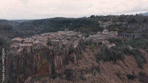Aerial view of Calcata Vecchia, an ancient medieval village perched on a rocky cliff in Lazio, during a gloomy day. Historic stone houses surrounded by forest and dramatic landscape of a scenic view.
