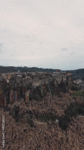 Aerial view of Calcata Vecchia, an ancient medieval village perched on a rocky cliff in Lazio, during a gloomy day. Historic stone houses surrounded by forest and dramatic landscape of a scenic view.

