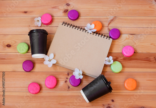 Notepad, brown paper coffee cups, colorful macaroons and apple tree flowers on wooden background. Top view with copy space.