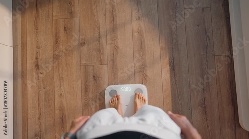 Top view of a young woman stepping onto a smart scale in a bright modern home. Morning routine moment focused on health, body tracking, and mindful weight management. Wooden floor and soft daylight
