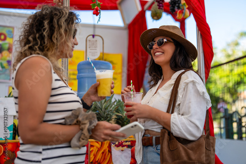 Quadro em tela Two smiling women friends enjoying fresh juice beverages at an outdoor street fo