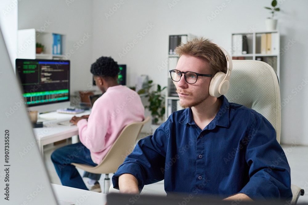 Obraz premium Young man wearing headphones working on computer at desk in office, Black young man sitting at adjacent workstation typing on keyboard, both focused on coding tasks