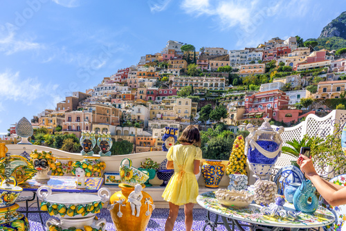 Amalfi Coast, Italy. Young girl in yellow dress admires colorful ceramic pottery on terrace overlooking hillside village of Positano, Italy with vibrant buildings and blue sky