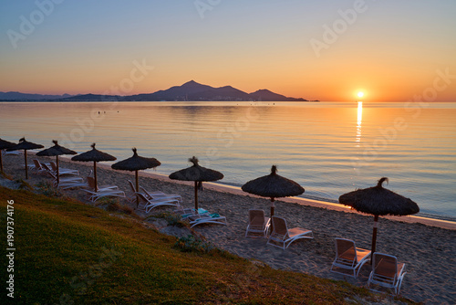 Breathtaking sunrise over Playa de Alcudia with straw parasols and sun loungers on the sandy beach, Bay of Alcudia, Balearic Islands, Spain.