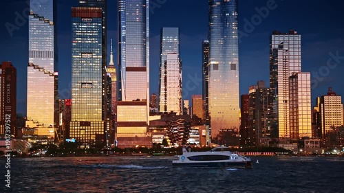 Bright soft light setting sun reflecting in the glass facades of New York skyscrapers. A modern boat moves along the waterfront with a Vessel Structure.