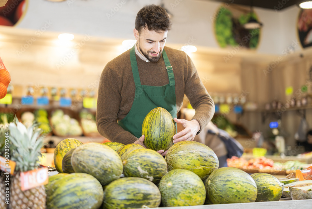 Fototapeta premium Eco food vegetable market, male worker and melon. Shop assistant add goods items in sales area. Worker add goods to showcase, sell imported and local farm agricultural products