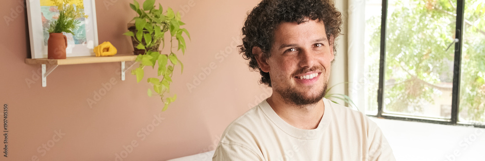 Obraz premium Young man with curly hair and beard crossing his arms and looking at the camera with smile while standing in cozy living room. Panoramic