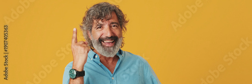 Elderly grey-haired bearded man wears blue shirt, thinks rubs chin comes up with ideas, raised finger, isolated on orange background in studio. Panoramic