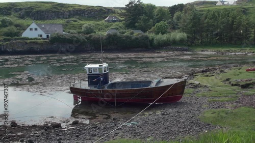 A boat rests on the land as the tide goes out. The sun shines on the bay with houses in the background and greenery surrounding the area.
