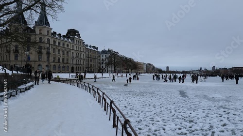 A view from a bridge onto a frozen canal in the city. Many people are walking on the ice. It's very cold and there's a lot of snow