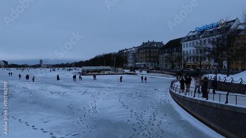 A view from a bridge onto a frozen canal in the city. Many people are walking on the ice. It's very cold and there's a lot of snow