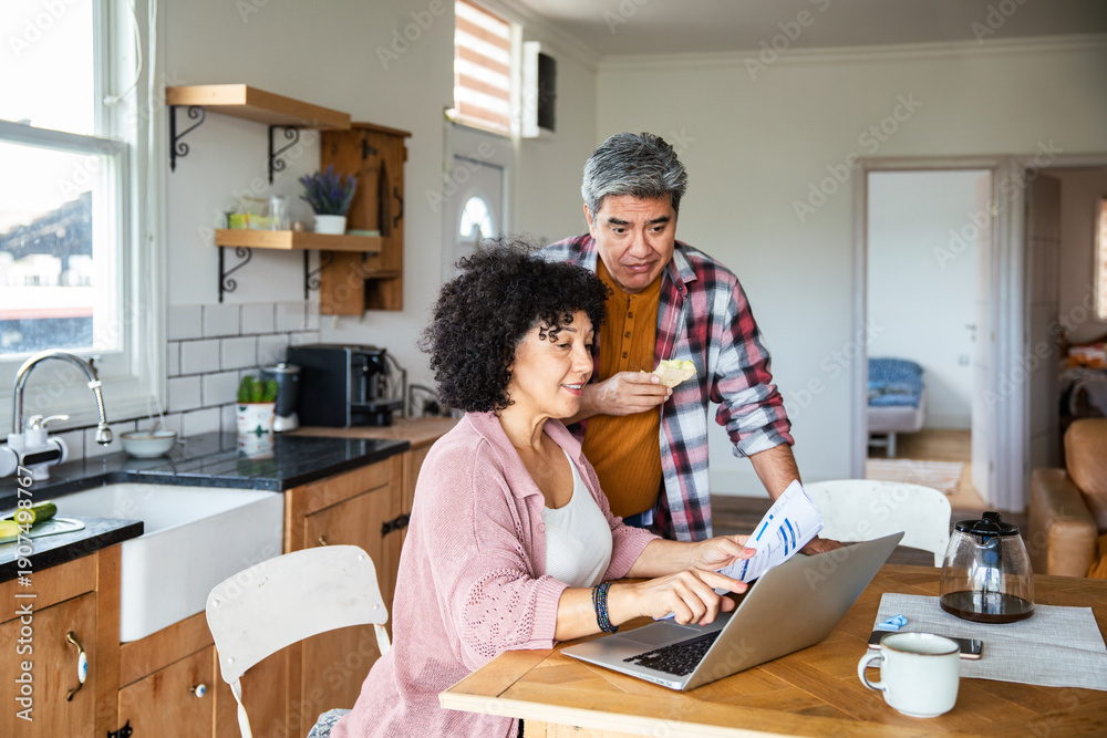 Obraz premium Middle-aged couple reviewing bills on laptop at kitchen table