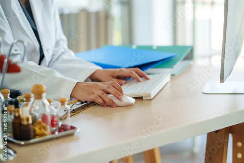 Calm healthcare office desk scene with doctor hands on keyboard and mouse modern computer medical folder clipboard glass bottles soft daylight clean workspace professional focus clinical setting