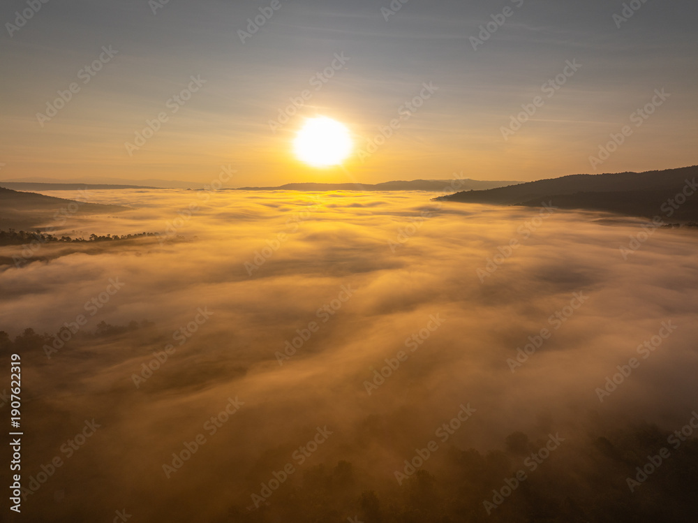 Fototapeta premium A cinematic drone aerial view of sunrise casting golden light over a misty mountain valley, with a sea of fog drifting through the landscape and distant peaks on the horizon.