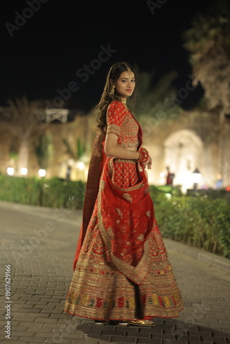 Beautiful young Indian woman dressed in an elegant red embroidered lehenga with traditional jewelry, posing outdoors at night