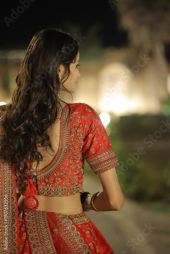 Beautiful young Indian woman dressed in an elegant red embroidered lehenga with traditional jewelry, posing outdoors at night