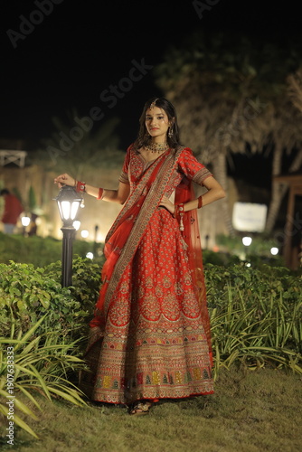 Beautiful young Indian woman dressed in an elegant red embroidered lehenga with traditional jewelry, posing outdoors at night