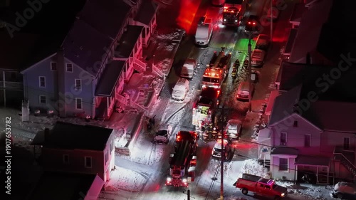 Firefighters responding to an emergency on snowy residential street in United States, with fire trucks, emergency lights, vehicles and houses illuminated during winter conditions. Aerial top down shot