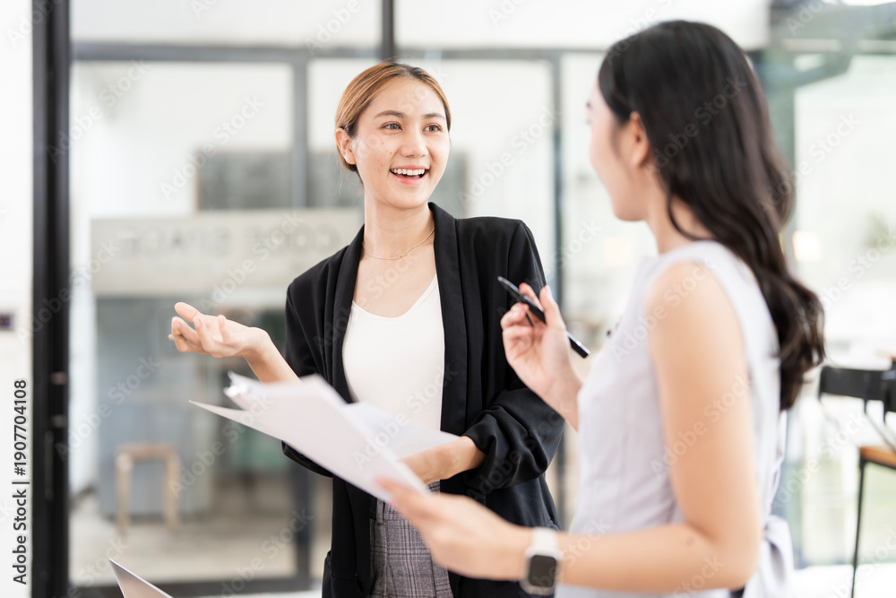 Obraz premium Two professional Asian businesswomen discussing project reports and collaborating in a bright, modern office environment, showcasing teamwork and effective communication.