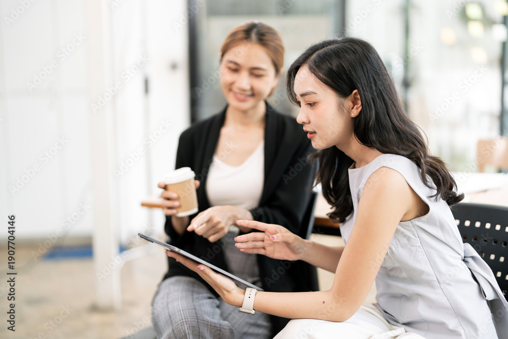 Fototapeta premium Two professional Asian businesswomen collaborating on a digital tablet and sharing ideas during a productive meeting in a modern office environment.