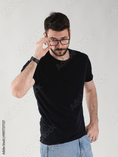 Handsome bearded confident young man in black t-shirt looking directly at camera with a focused expression while adjusting his eyeglasses with one hand.