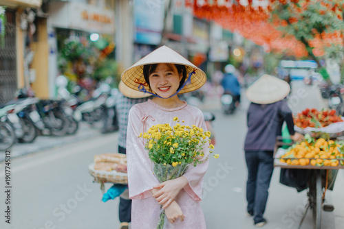 A smiling young woman in a traditional conical hat holds yellow flowers on a street in Old Quarter, Hanoi, Vietnam. She is surrounded by vendors and motorbikes.