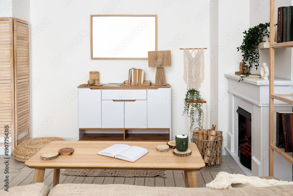 Naklejka premium Interior of living room with commode, coffee table, houseplants, fireplace and firewood in wicker basket near white wall