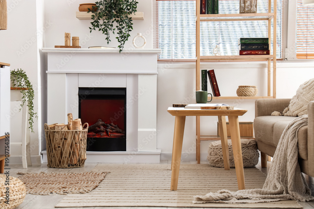 Naklejka premium Interior of living room with sofa, shelving unit, coffee table, fireplace, houseplants and firewood in wicker basket