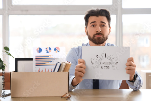 Fired young man holding paper with text I NEED A JOB at table in office
